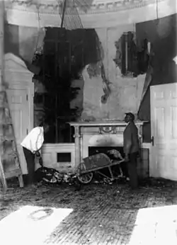 a black and white image of a room ravaged by fire with a man holding a wheelbarrow full of debris in front of a fireplace