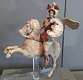 A female warrior is seated on the back of a rearing horse. She wears a one-shouldered tunic that flows behind her, a feathered helmet, a shield and jewelry, which are decorated with reds, browns, white, and gold. The horse's bridle and reins are similarly decorated.