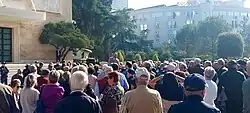 Një grup njerëzish para godinës së Kryeministrisë së Rep. së Shqipërisë dhe Arlind Qori duke ligjëruar.