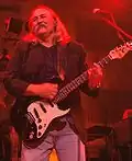 A man with a mustache and shoulder-length hair wearing jeans and holding a guitar, standing behind a microphone.