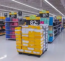 Several people shopping in an area with high shelves on the right stacked with spiral notebooks and other stationery products in open yellow boxes. At the top of the shelves are several blue signs with a small stylized starburst logo in yellow and "Everyday Low Price" in white text, on a red background. Strip fluorescent lights on the ceiling illuminate the scene; a yellow sign hanging from the ceiling has an octagon with "back to school" and text in English and Spanish beneath it. On the left are shelves reaching camera height; a sign in the front bottom says "$9.97".