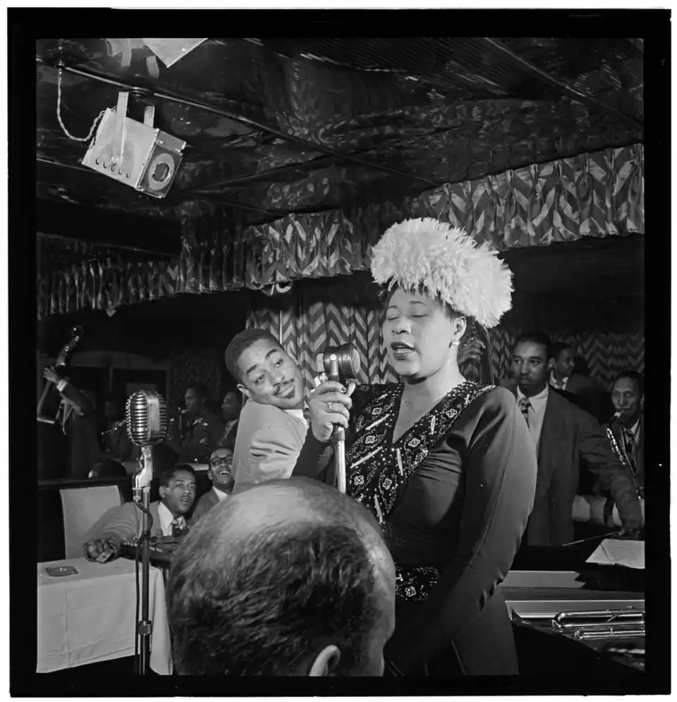 Fitzegeral is singing with a microphone inside a jazz club. Ray Brown stands behind her; Dizzy Gillespie and Milt Jackson sit in a table in the background; and Timme Roesenkratz in the foreground.