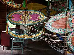 A wau-maker's workshop. Foreground: Wau frames. Background: A completed wau. Further background: The wau-maker.