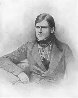 Black and white image of a seated man from the waist up. He is dressed in a nineteenth century suit, clean shaven and looking to his right.