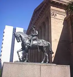 Equestrian statue in dark grey metal of George&nbsp;V in military dress uniform on a plinth of red granite outside a Classical building of red sandstone