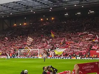 A single tiered stand that contains thousands of people. Several flags are being waved. In front of the stand is a grass pitch with a goal.