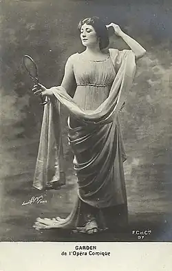 Portrait of a young white woman with dark hair, standing draped in a classical Roman gown and sandals, admiring herself in a handheld mirror