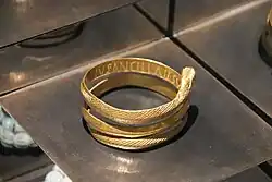 A large gold bracelet that coils 2-3 times around the wrist is in this photograph. It is behind a display case perhaps at a museum. The bracelet has patterns on it including snakeskin, feather, and a section that is patterned. It contains about a pound of gold.