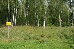 A cleared strip of land in the middle of a wooded area. In the center are two small wooden posts, one painted red and white and the other green and red. On either side are signs in Polish stating that this is the international border and that crossing it here is prohibited
