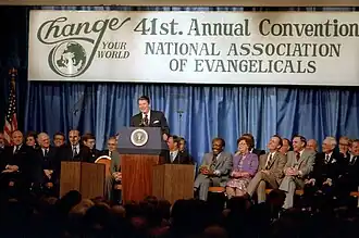 Reagan, smiling, standing behind a lectern with the presidential seal and a microphone. Behind him is a banner for the convention, with the slogan "Change Your World" and the title "41st. Annual Convention NATIONAL ASSOCIATION OF EVANGELICALS". Behind the banner are curtains. In front of the banner and curtains but behind Reagan are two rows of seated people, about 20 total. Most seem in good spirits with smiles and expressiveness. Most are Caucasian men. Two women are visible, as is a Black man.