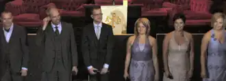 Six people standing in a row, three men and three women, the men wearing black suits with white shirts and the women wearing dresses