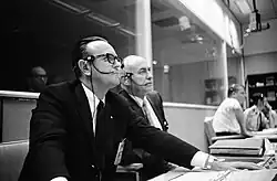 Two middle-aged men sit at consoles, wearing headsets