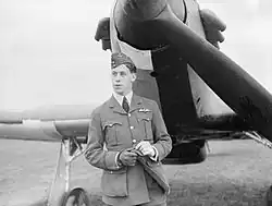 A black and white photograph of the head and torso of a man in uniform, wearing a side cap and gloves, standing in front of a fighter aircraft