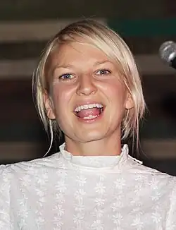 A woman with a fair complexion and short, bleached hair wearing a modest white top looks into the camera, mouth open; a microphone is seen at the edge of the photo.