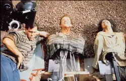 A photograph of animatronics of Arnold Schwarzenegger and Rachel Tictoin, built from the chest upwards, lying down next to a crew member