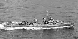 A ship plows through the water. It has two guns forward, and two aft.