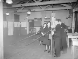 A wargame in progress. The men on the far-right command the escort ships and may only view the game board through holes in the screen. The woman in the foreground is Jean Laidlaw.